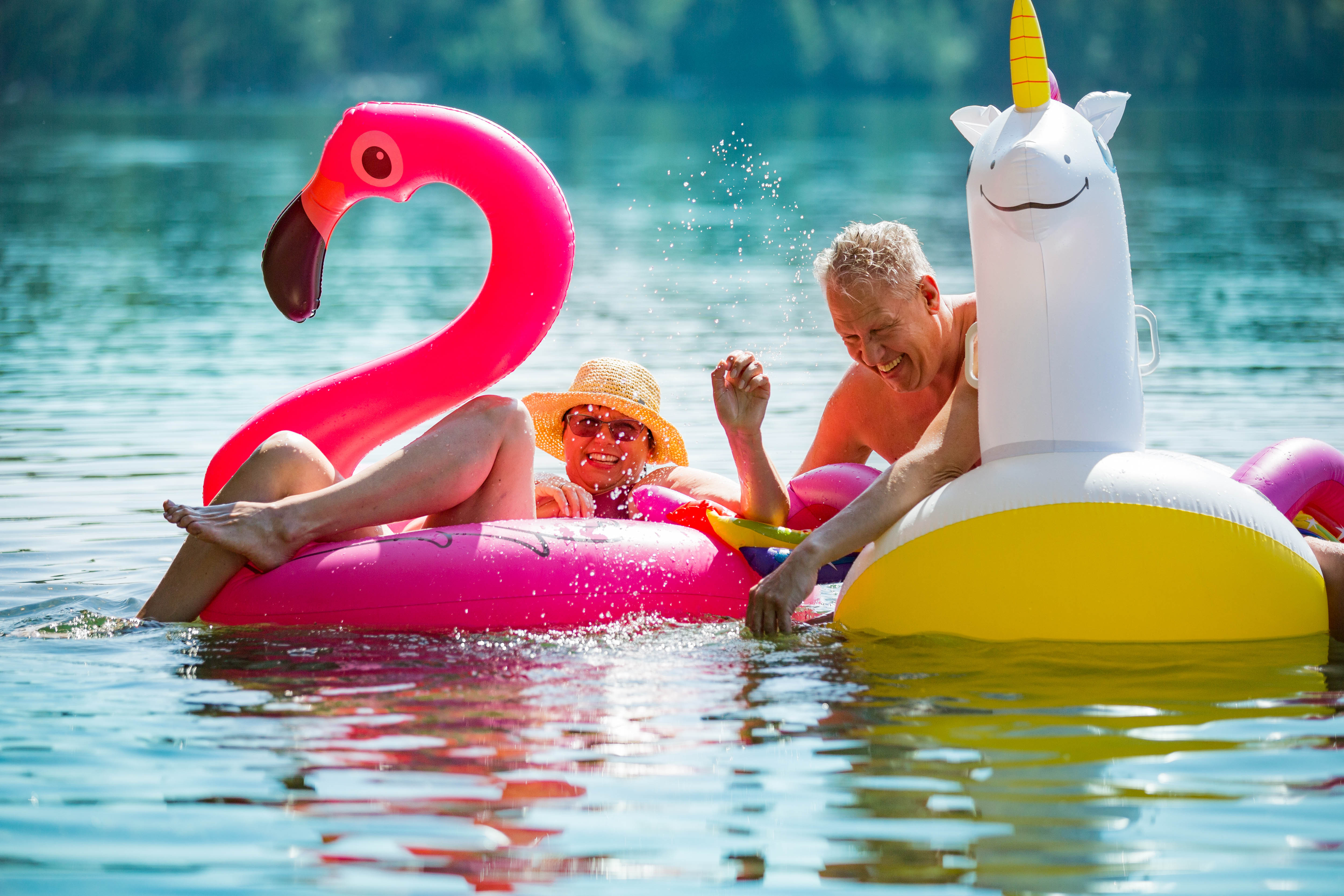 Older man and woman floating on flamingo and unicorn floaties in a lake Older man and woman floating on flamingo and unicorn floaties in a lake